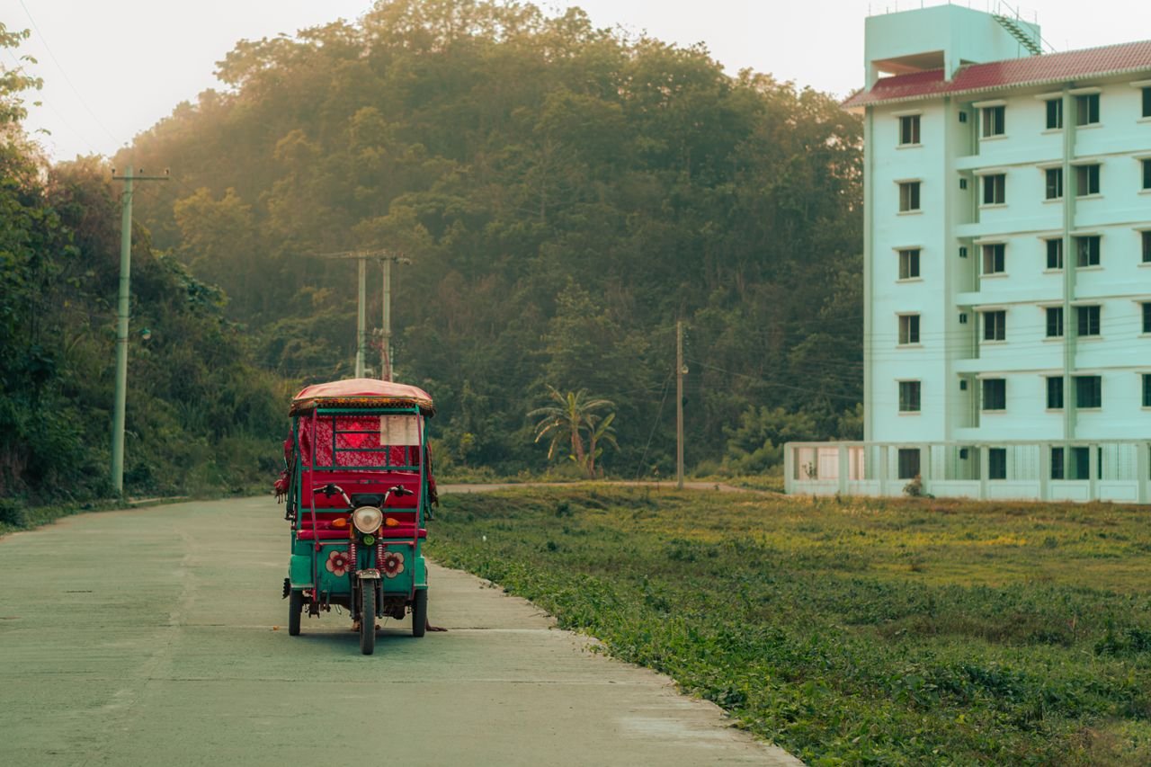 About A vibrant rickshaw travels down a rural road in Chittagong, Bangladesh, near a building and lush greenery.