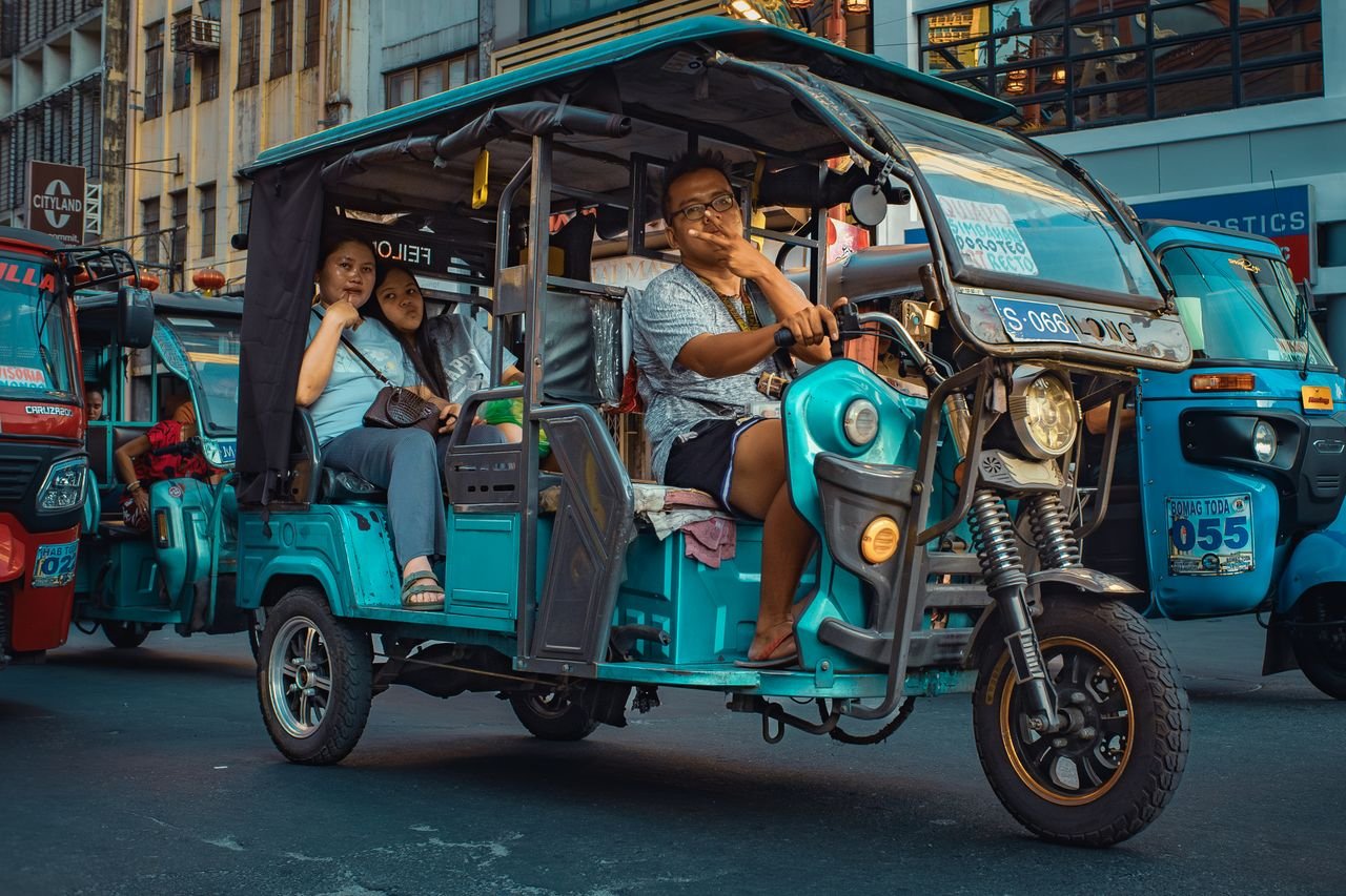 Services Three people riding a turquoise tricycle on bustling Manila street during the day.